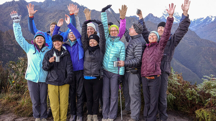 A group of eleven people wearing outdoor gear stand together, some with arms raised, against a backdrop of mountains. They appear cheerful and some hold cups, showcasing their hiking essentials for the Inca Trail.