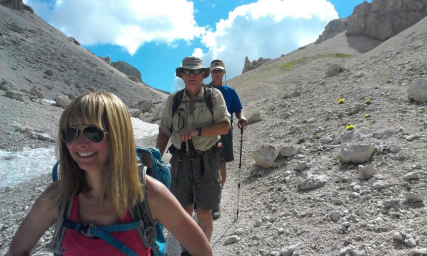 Three hikers equipped with backpacks and hiking poles traverse a rocky mountainous trail under a partly cloudy sky. The lead hiker, Tania, wears sunglasses with the other two following close behind.