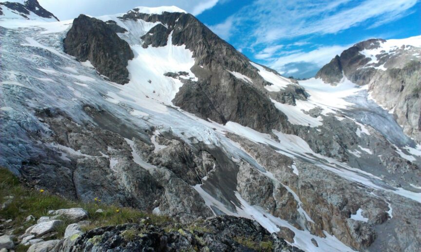 Snow-covered rocky mountains under a blue sky with clouds, featuring patches of snow and ice on the rugged terrain. Tania marveled at the breathtaking view, contemplating her next move along the winding trail.