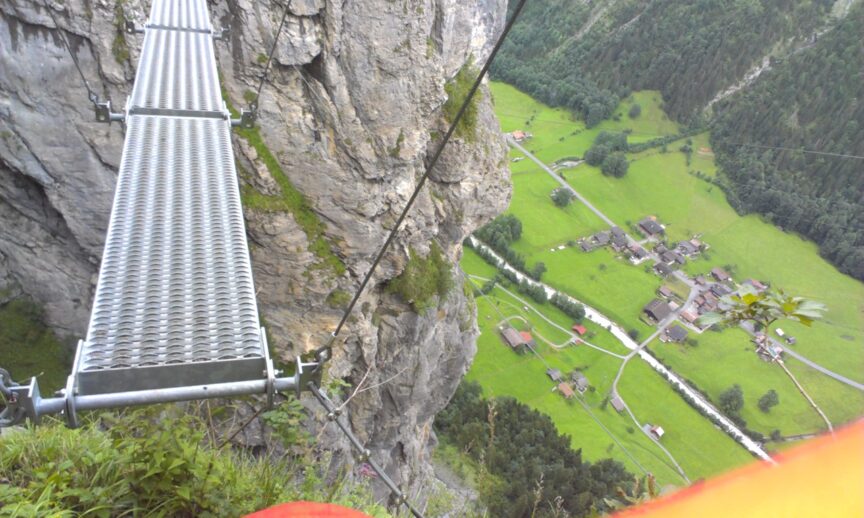 A narrow metal bridge is suspended between two rocky cliffs, high above a green valley with scattered buildings and trees, offering the perfect setting for an outdoor adventure on Tania's trail.