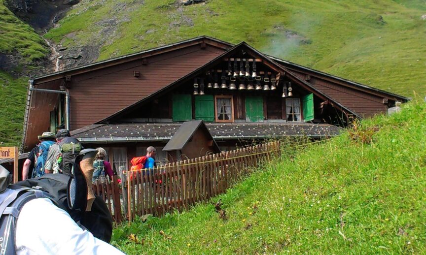 Hikers approach a wooden mountain hut with green shutters. Cowbells line the eaves of the roof as Tania leads them along the trail. The hut is situated on a grassy hillside.
