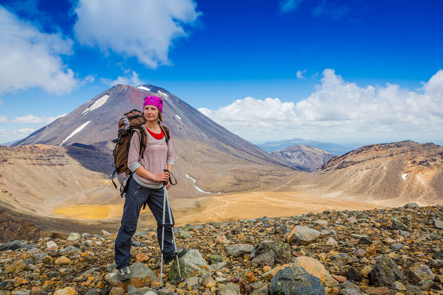 A person with hiking gear stands on a rocky terrain, with iconic peaks and a mountain framed against a clear blue sky in the background.