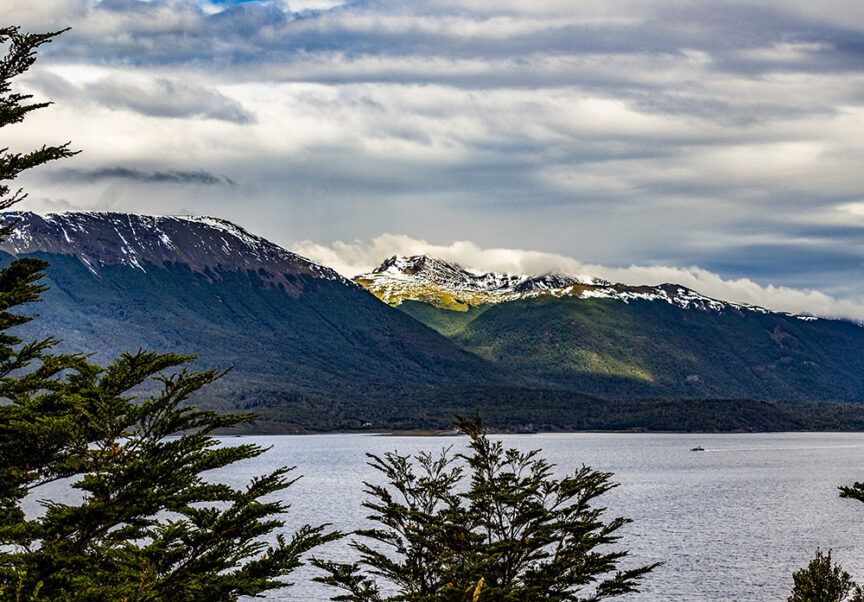 Mountain range with patches of snow under a cloudy sky, overlooking a body of water with trees in the foreground, evoking the wild beauty of Ushuaia. This "End of the World" vista promises adventures waiting to be discovered.