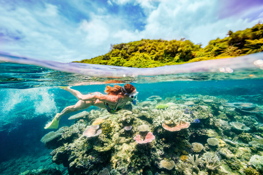 A snorkeler swims underwater over vibrant coral reefs with an island and blue sky in the background, showcasing the magic of snorkeling.