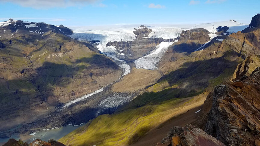 A rugged mountain landscape with a glacier winding through the valleys. Snow-capped peaks and patches of green vegetation are visible under a partly cloudy sky, perfect for scenic hikes in Iceland. At night, you might even catch the Northern Lights dancing above.