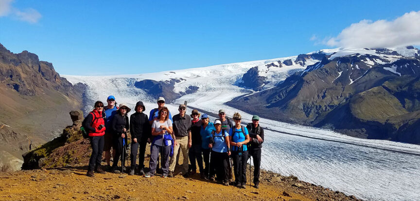 A group of people stand together on rocky terrain in Iceland, with a vast glacier and mountainous landscape in the background under a clear blue sky, after one of their exhilarating hikes.