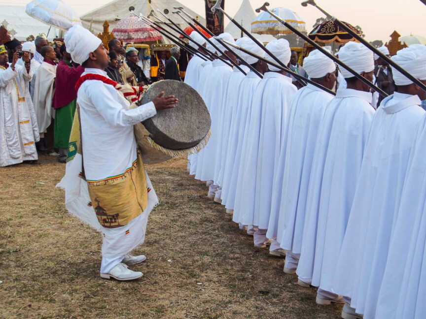 A group of people dressed in white traditional garments perform in a colorful ceremonial event in Ethiopia. One person in the foreground plays a large drum while others hold long thin instruments upright.