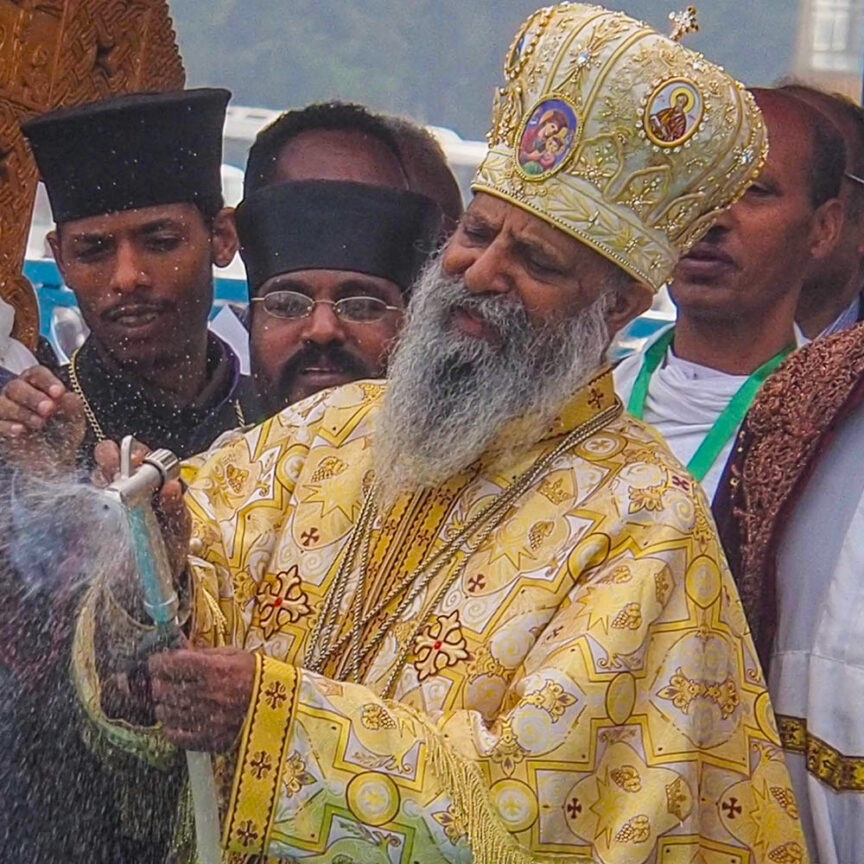 A bearded man in ornate, colorful religious attire and a gold crown sprinkles water during a ceremonial event in Ethiopia, surrounded by other men in black and white garments.
