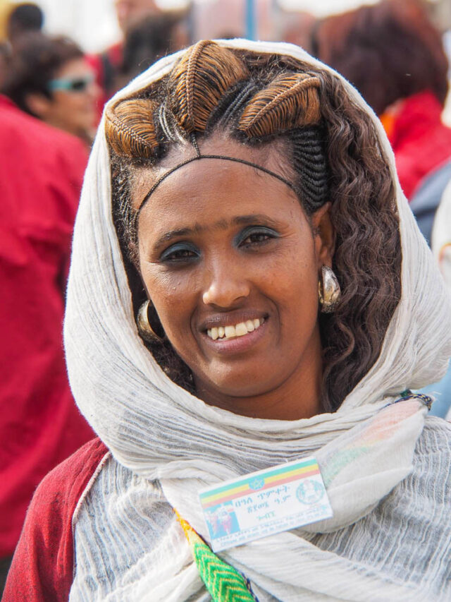 A woman with braided hair and a white shawl smiles. She wears a name tag, embodying the vibrant spirit of Ethiopian tourism, and is surrounded by people in colorful red clothing.
