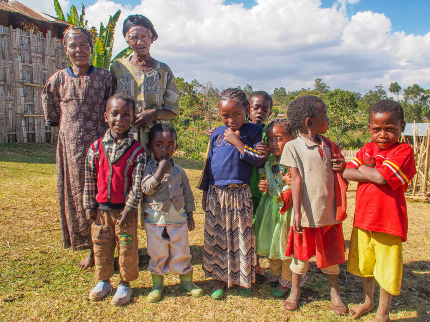 A group of nine children and one elderly woman stand together outdoors, posing for the photo in Ethiopia. The background features lush greenery, trees, and a partly cloudy sky, while their colorful clothing adds vibrancy to the scene.