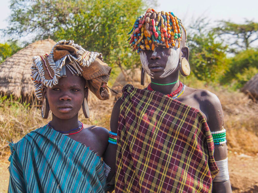 Two people in colorful traditional attire, standing outdoors in Colorful Ethiopia. One wears a beaded headpiece; both have body paint and jewelry. Huts and lush vegetation are visible in the background.