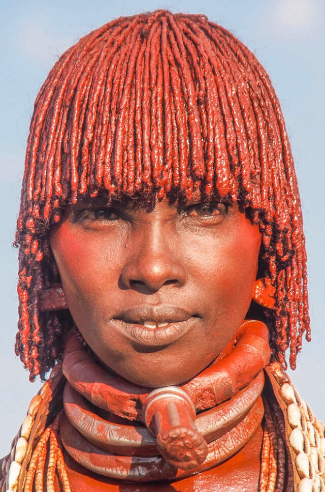 A portrait of a person with intricate red braids, adorned in colorful traditional attire and accessories, looking directly at the camera; this image captures the vibrant essence of Ethiopia, inviting viewers to travel and explore its rich cultural heritage.