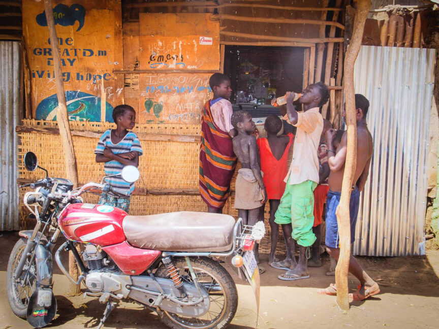 A group of children stands outside a small shop with corrugated metal walls and a woven facade. A colorful red motorcycle is parked nearby. A painted sign, reminiscent of Ethiopia's vibrant streets, is partially visible above the shop entrance.