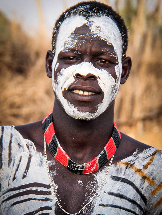 A man with white and black body paint on his face and chest, wearing a red and black beaded necklace and a gold chain, smiles while standing outdoors in Ethiopia. His colorful appearance highlights the vibrant culture of the region.