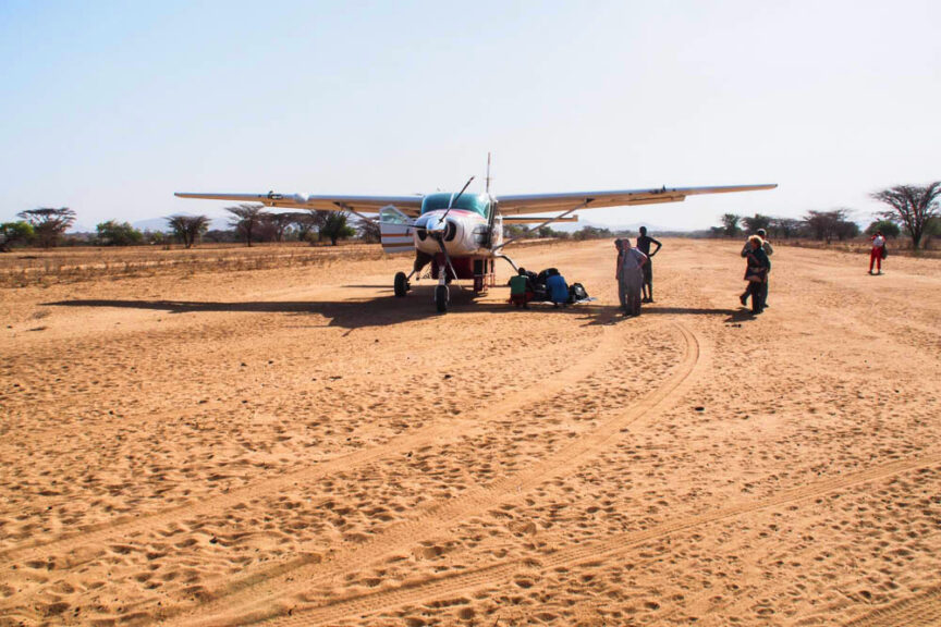 A small propeller plane is parked on a dirt airstrip in Ethiopia, with several people standing around it, some handling colorful luggage. The surrounding area is dry and sandy, with scattered trees.