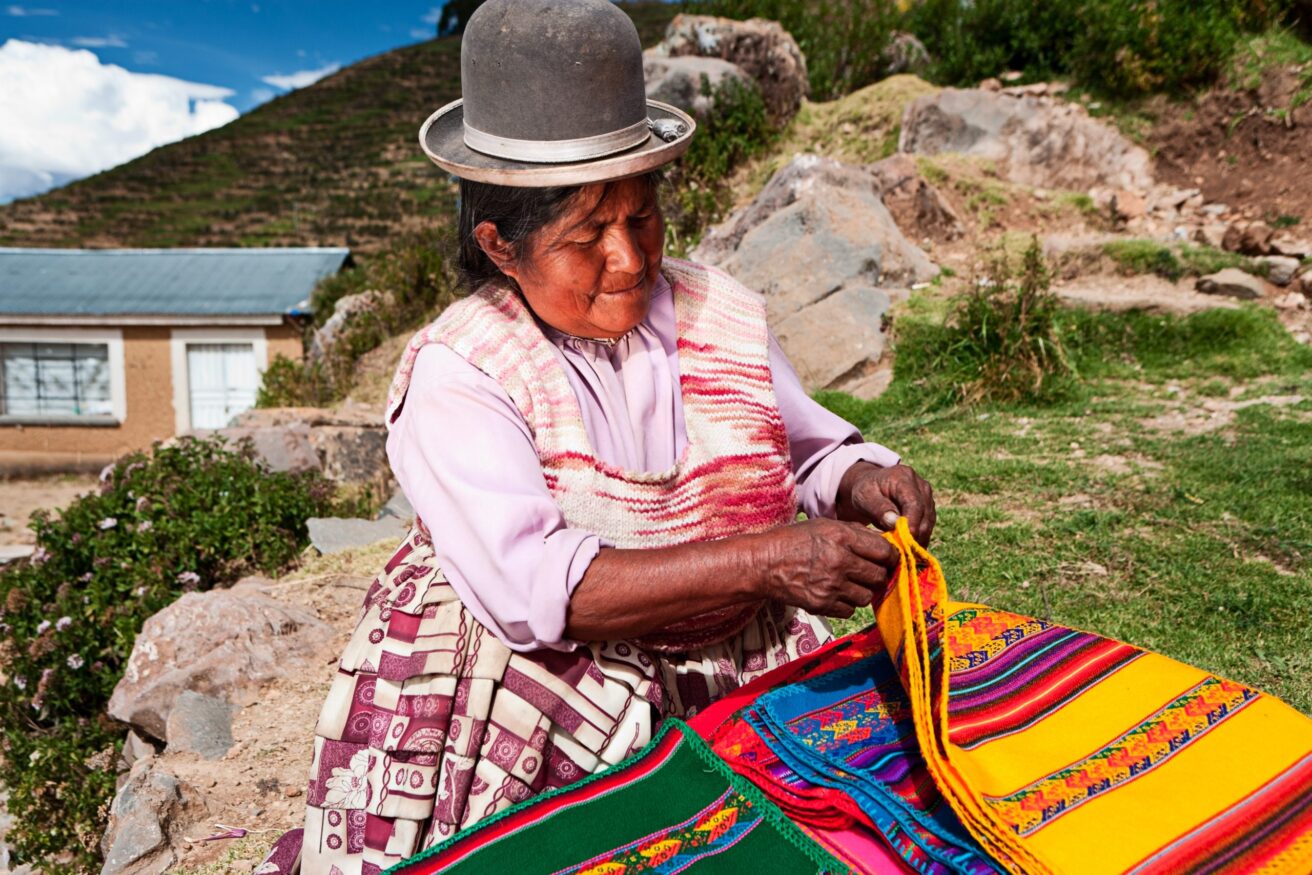 An elderly woman wearing traditional clothing, including a hat, arranges colorful woven textiles outdoors, with greenery and a building in the background. This scene showcases the power of tourism in promoting sustainable travel and supporting community-based tourism initiatives.