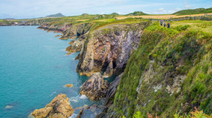Cliffside view of a coastal landscape in Wales with rocky cliffs, lush greenery, and the blue ocean. A group of people enjoys hiking on a path, experiencing the refreshing breeze under a Lark Full Cloud sky.