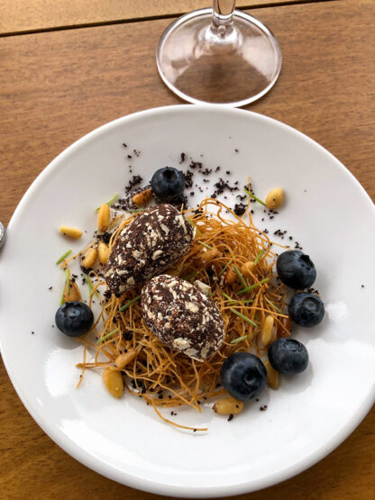 A gourmet dessert featuring two chocolate truffles, crispy noodles, blueberries, pine nuts, and green herbs on a white plate is presented as if ready for a refined evening on a Small Ship cruising Alaska's Inside Passage, with an empty wine glass in the background.