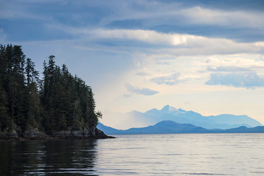 Calm water with a dense forested shoreline on the left and distant mountain range under a cloudy sky, reminiscent of Alaska's Inside Passage near Sitka.