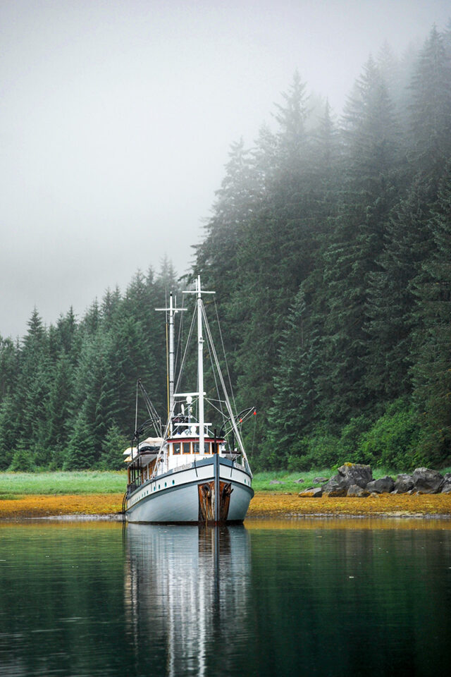 A small ship is anchored on calm water, surrounded by dense, fog-covered evergreen forest in the Sitka region of the Alaska Inside Passage.