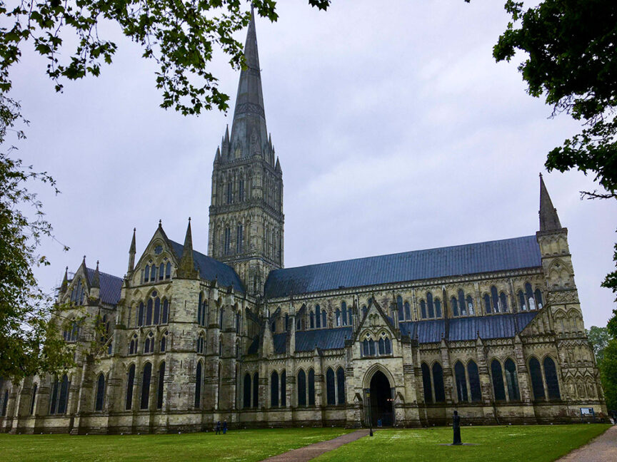 An exterior view of Salisbury Cathedral with its towering spire, Gothic architecture, and lawn in the foreground under a cloudy sky—truly a sight from Britain's Land Lost in Time.