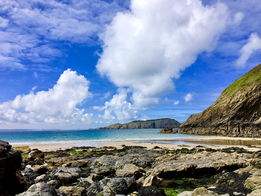 A rocky shoreline with patches of greenery, seemingly lost in time, leads to a sandy beach and turquoise ocean, with cloudy blue skies and hills in the background.