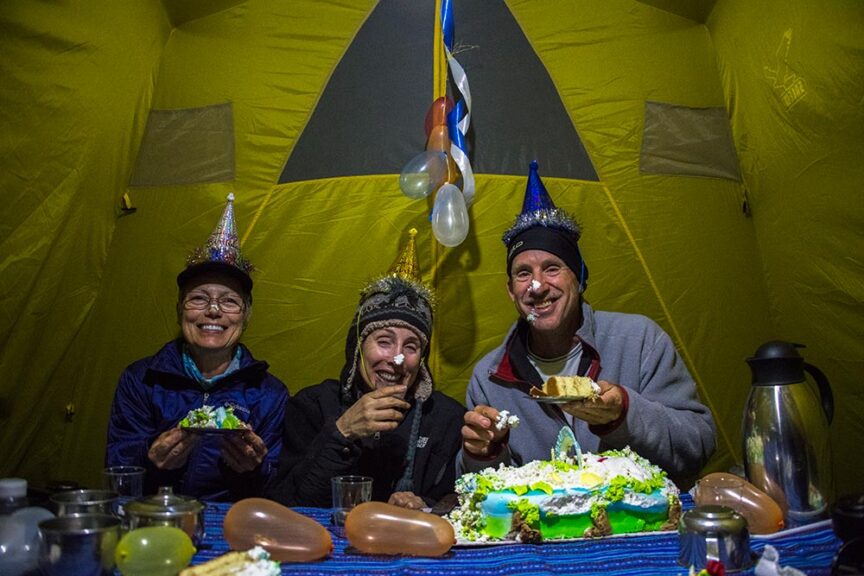 Three people wearing party hats are inside a tent, celebrating a 60th birthday with a cake. They appear to be enjoying the festivities, with some frosting on their faces. Party balloons are hung behind them as they revel in the wilderness of Peru.
