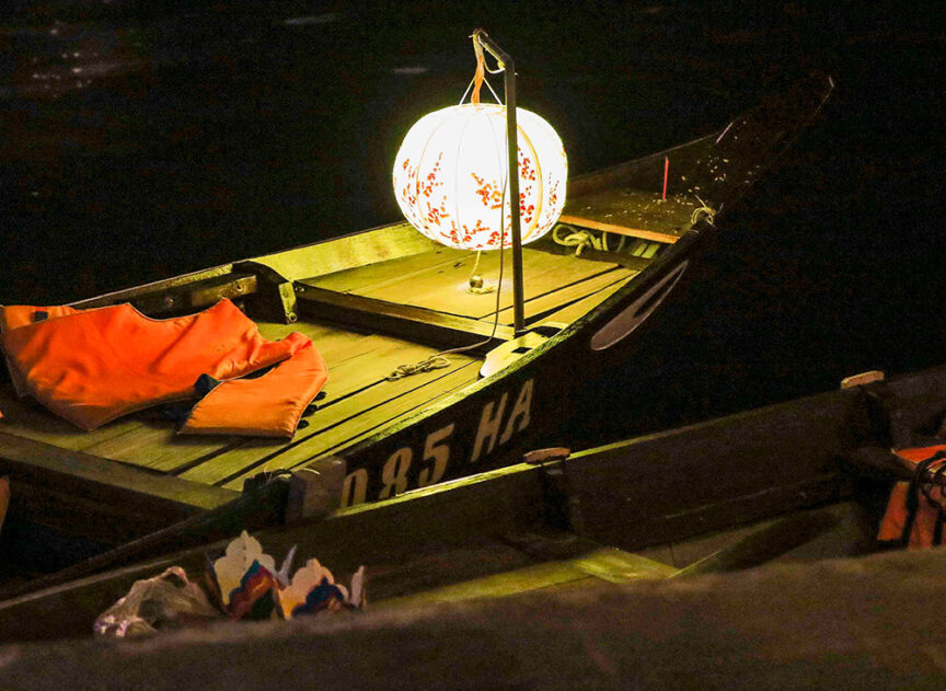 A small wooden boat with the registration number "085 HA" is anchored at night, its charm reminiscent of an "Adventures in Indochine" scene. Illuminated by a hanging lantern, the boat's seats are adorned with orange life jackets, creating a warm and inviting ambiance.