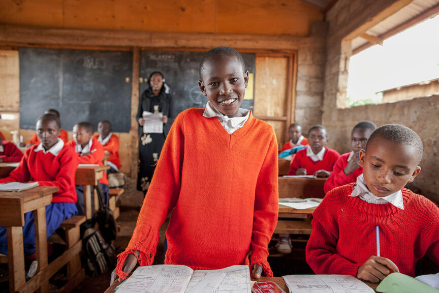 Children in a classroom, some sitting at desks writing in notebooks and one standing and smiling at the camera. They are wearing bright red sweaters and the classroom has wooden furniture and a blackboard. Despite the challenges of the pandemic, they radiate joy, reminding us why we should donate to support their education.