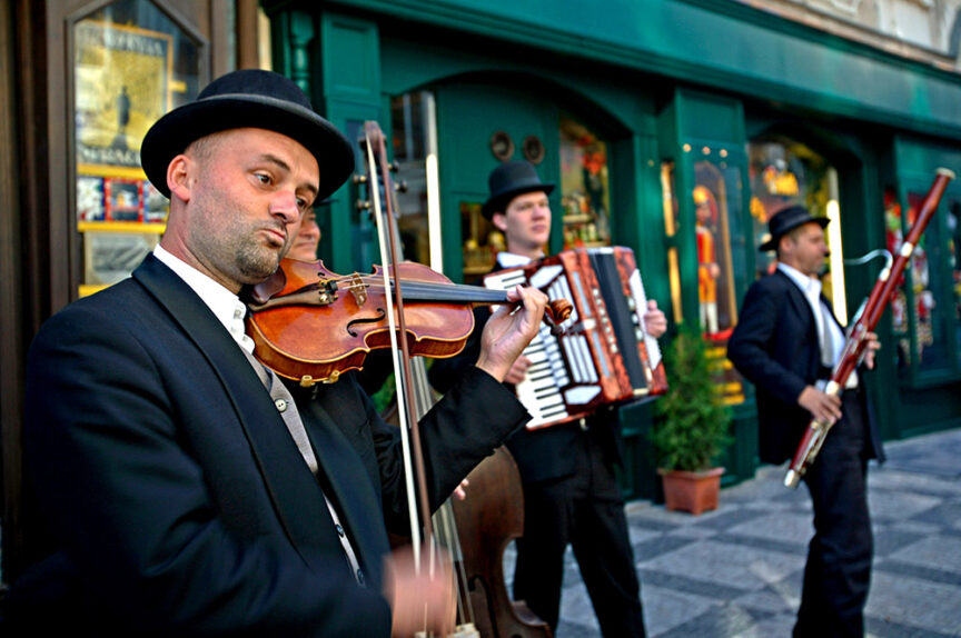 Four musicians in formal attire play instruments (violin, accordion, bassoon) on a city street, delighting types of travelers exploring Europe with their melodies as they stand in front of green storefronts.
