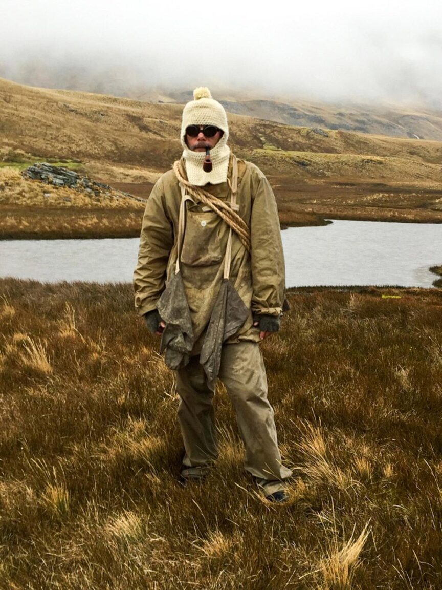 A person dressed in heavy outdoor clothing and a knit hat stands in a grassy, hilly landscape near a body of water under a cloudy sky, embarking on the journey to explore the Seventh Continent.