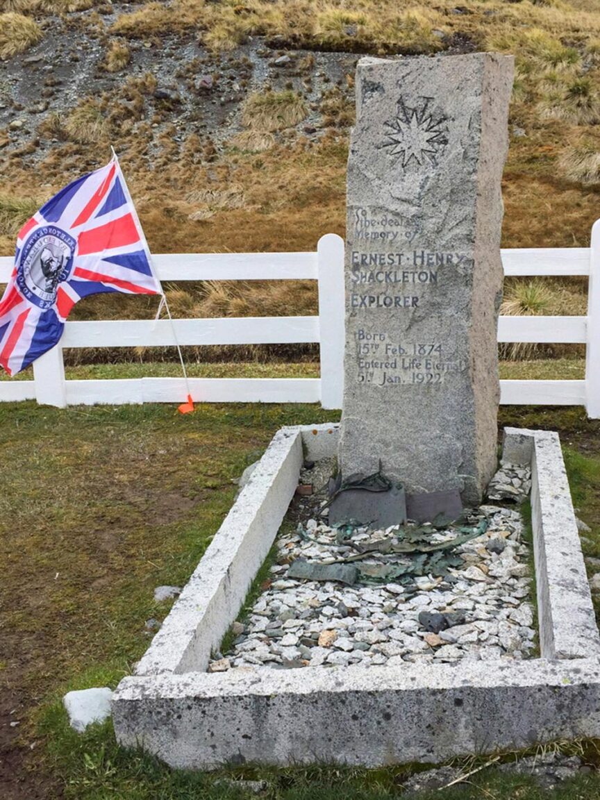 A gravestone and British flag mark the resting place of explorer Ernest Henry Shackleton, born February 15, 1874, and died January 5, 1922. Situated in a fenced area with rocky terrain, this site commemorates his adventurous journey to the Seventh Continent.