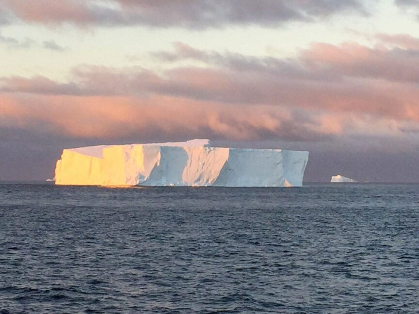 A large iceberg floats in calm ocean waters under a sky with patches of clouds tinged with pink hues from the setting or rising sun, marking the start of an adventure to the mysterious Seventh Continent.