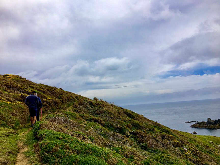 Person hiking on a narrow dirt path along a grassy, hilly coastline with the sea and cloudy sky in the background, experiencing Britain's Land Lost in Time.