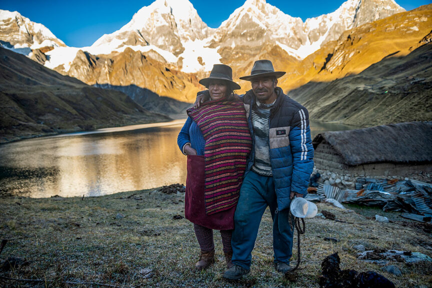 Two people stand side by side in front of a tranquil lake with snow-capped mountains in the background, both wearing hats and warm clothing. A thatched-roof structure is visible near the lake, set against the majestic peaks of the Cordillera Huayhuash.