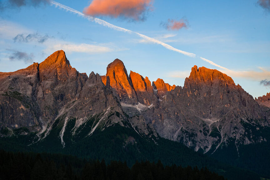 Mountain range with rugged peaks illuminated by the setting sun, contrasting against a clear sky with a single white contrail. Dark forested area lies at the base of the mountains—an inviting scene for exploring the Great Dolomites on your next travel adventure.