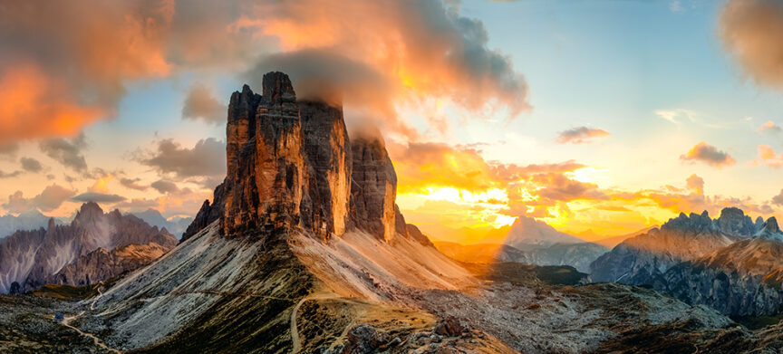 Panoramic view of the iconic Tre Cime di Lavaredo peaks in the Dolomites at sunset, with dramatic clouds and vibrant sky hues, reminiscent of a world steeped in legends.