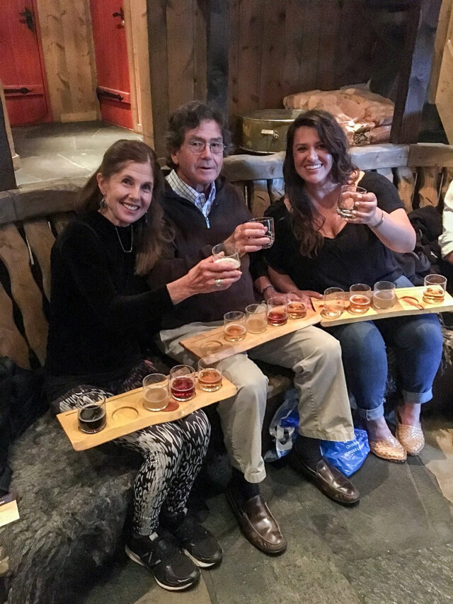 Three people, seated on a bench and smiling, raise their beer flights in a toast. Against the stunning backdrop of Norway's Fjord Country, they relish the simple delights of the moment, each sip as refreshing as their recent hikes.