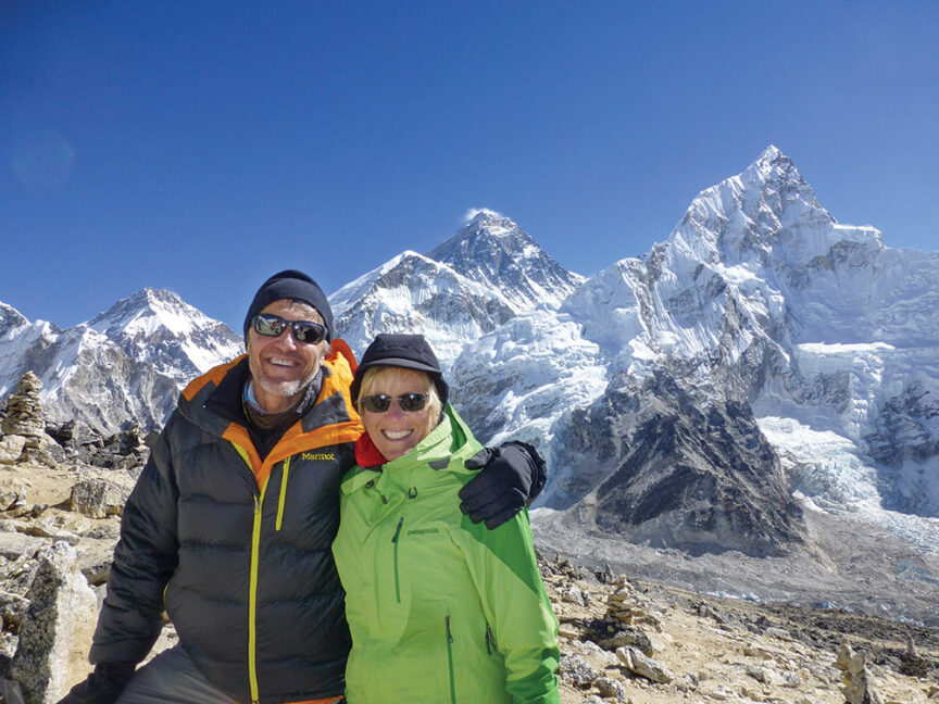 Two people in winter gear stand in front of iconic peaks, their silhouettes framed against the clear blue skies and snow-covered mountains.