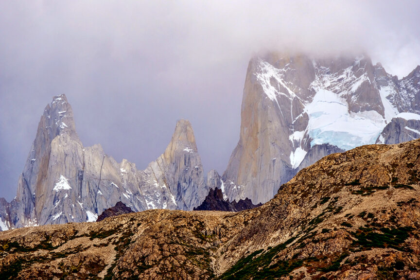 A rugged mountain range with sharp peaks partially covered in snow beneath a cloudy sky, reminiscent of Patagonia, with rocky terrain and sparse vegetation in the foreground, leaving lasting impressions.
