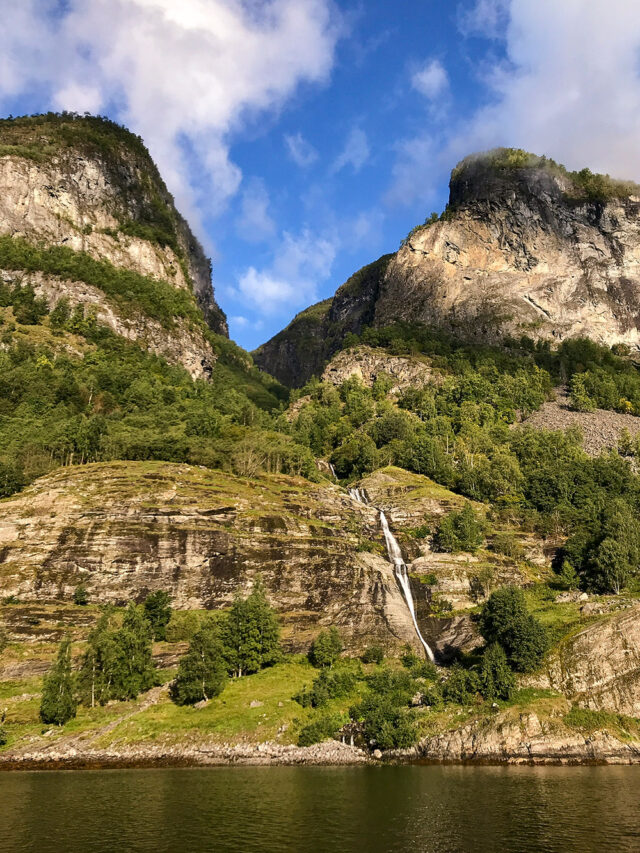 A narrow waterfall cascades down a rocky, green mountainside under a partly cloudy blue sky, with a calm fjord in the foreground—a perfect scene for hikes in Norway.