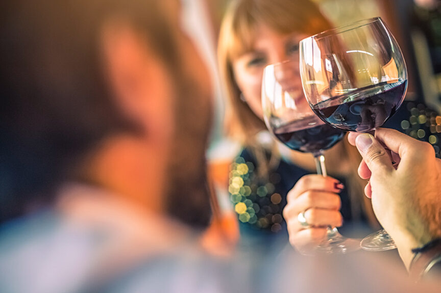 Two people clink glasses of red wine, smiling and facing each other in a warmly lit setting, embarking on a delicious culinary journey of the senses.