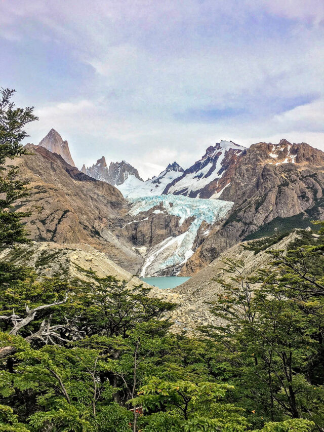 Snow-capped mountains and glaciers form a stunning backdrop to the rocky landscape and green shrubs in the foreground, embodying the epic hikes that Patagonia's national parks are renowned for.