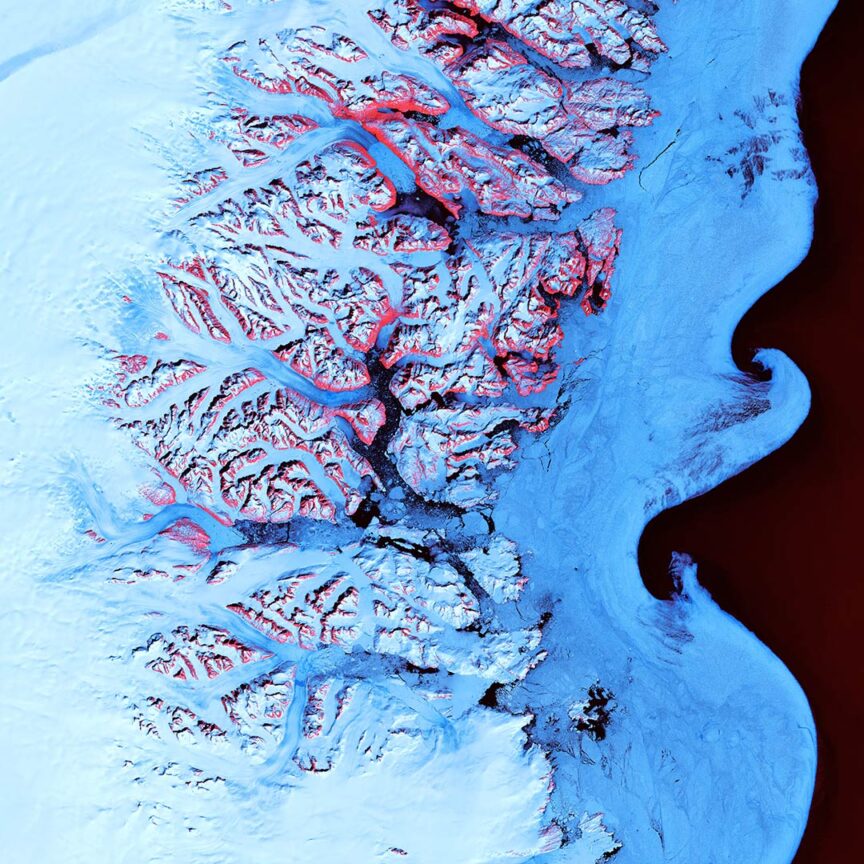 A stunning aerial view of a glacier with bright blue ice and red rivers flowing through crevasses, alongside a dark water body, showcases the Earth's breathtaking landscapes.
