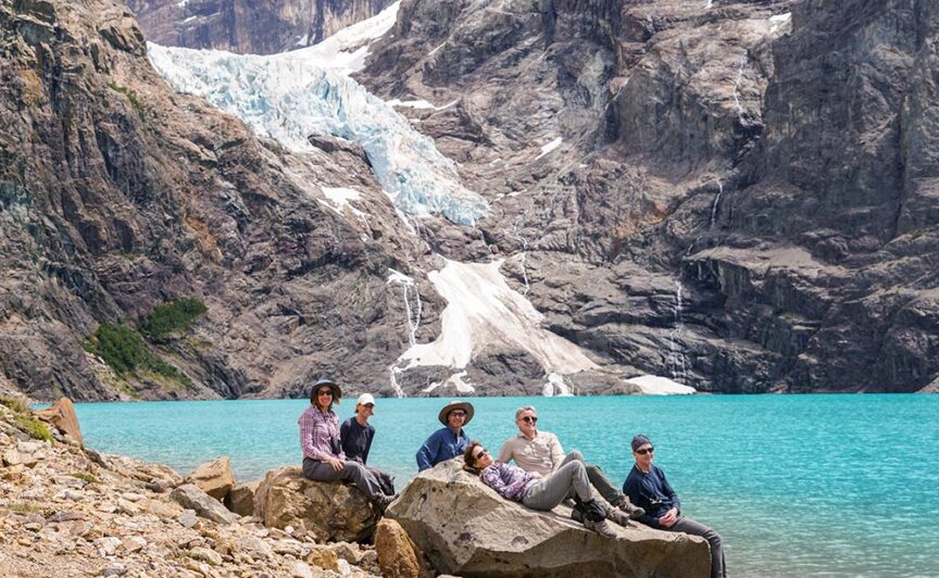 A group of six people in outdoor attire sit and recline on rocks by a turquoise lake, with snowy mountains in the background, embodying the spirit of Patagonia.