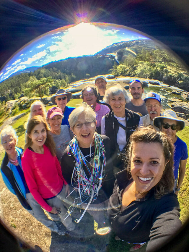 A group of people smiling and posing for a selfie outdoors with a fisheye lens effect. They are surrounded by the greenery and mountains of Norway under a sunny, blue sky, capturing an idyllic moment in Fjord Country.
