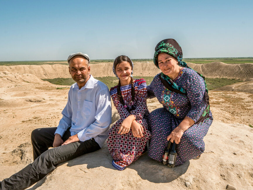 A man, a girl, and a woman, all dressed in traditional clothing, sit on a rocky landscape with a view of rolling hills under a clear blue sky, embodying the essence of a cultural journey along the historic Silk Road.