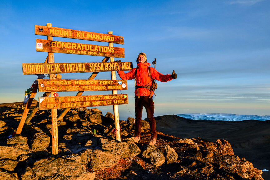 A person stands next to signs at the summit of Mount Kilimanjaro, indicating it's the highest point in Africa, at an elevation of 5895 meters above sea level. For those interested in this adventure, remember that trips to book should be planned well in advance.