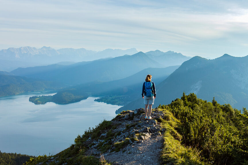 A person with a pack stands on a rocky outcrop overlooking a large lake and distant mountains covered in greenery under a blue sky, embodying the spirit of Alps hiking and adventure.
