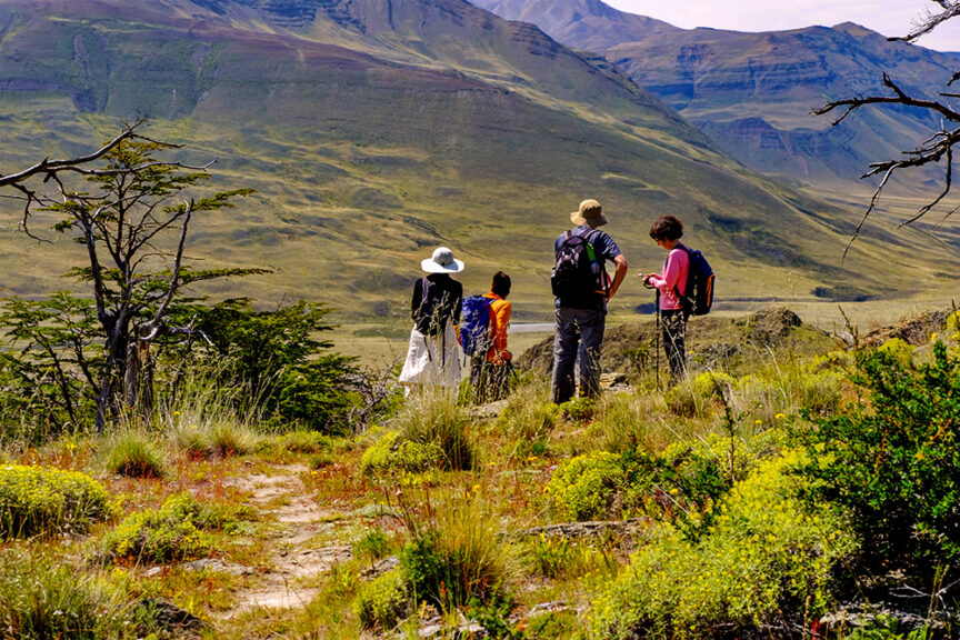 Four people with backpacks stand on a hiking trail, overlooking a mountainous landscape with sparse vegetation and greenery, capturing the essence of Patagonia impressions.